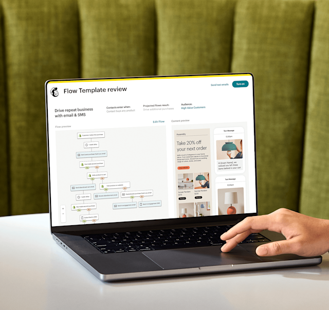 Image of a person sitting at a desk, looking at a laptop screen displaying Mailchimp’s Flow Template review feature. The feature shows a preview of an automation flow and an abstract view of email and SMS interfaces from the fictitious brand Assembly.